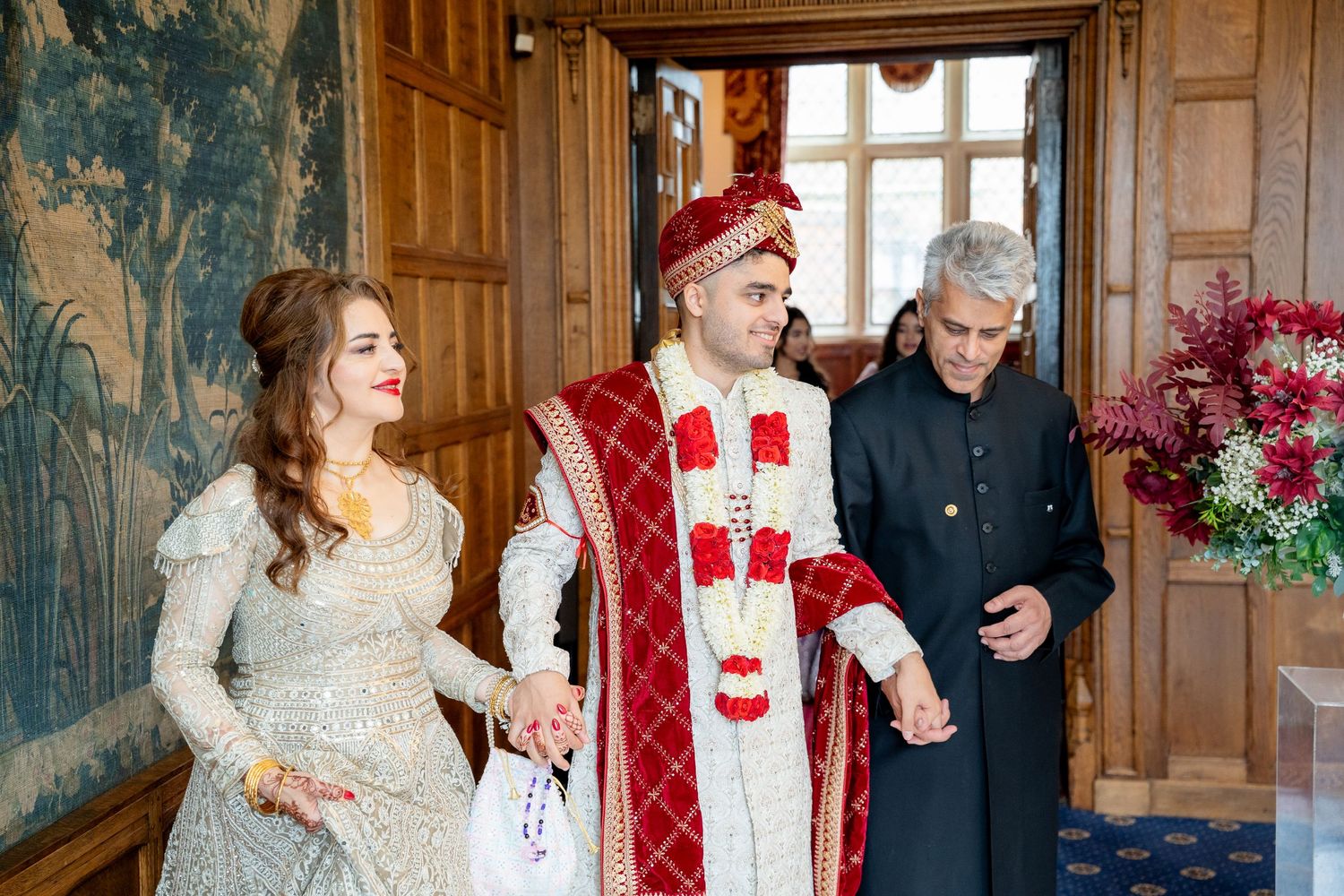 South Asian groom in red sherwani and bride in silver lehenga enter wedding venue with elder guest.