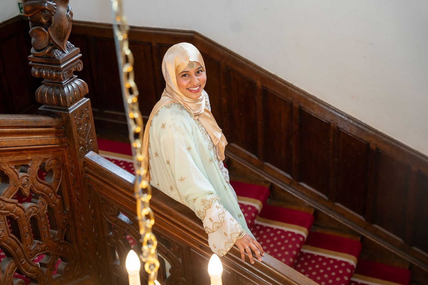 Woman in elegant light green hijab and embroidered outfit smiling on ornate wooden staircase.