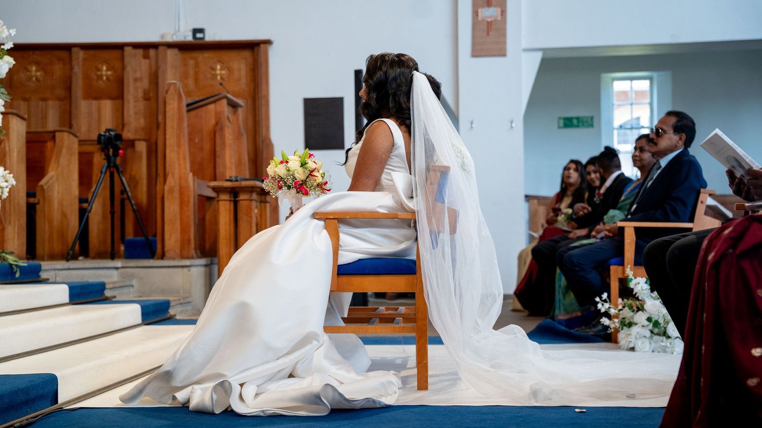 Bride in a white gown and long veil seated at the altar during a church wedding ceremony.