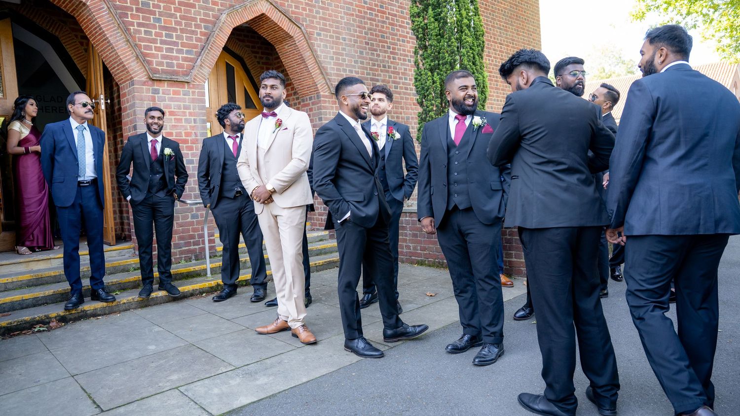Groomsmen in black suits gather outside brick church before wedding ceremony on sunny day.