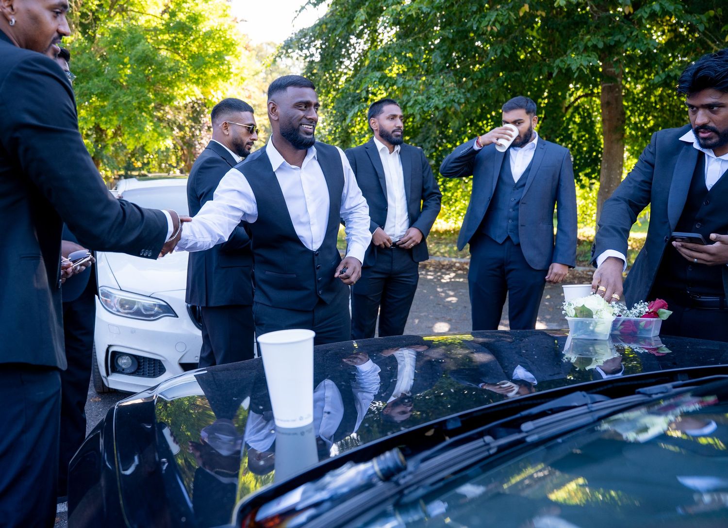 Groomsmen in black suits gather around a decorated car outdoors on a sunny day.