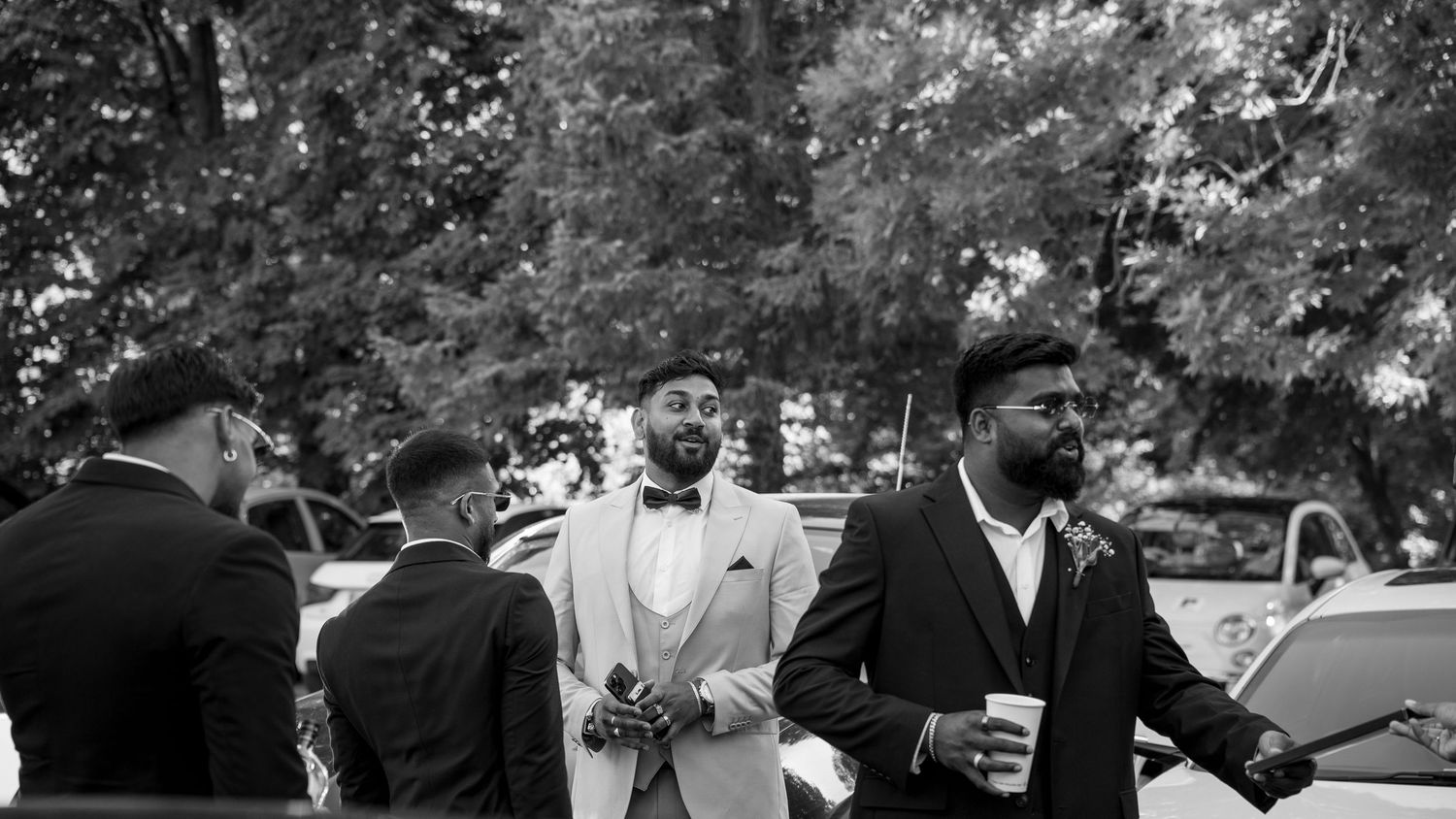 Black and white candid photo of groom in bow tie with groomsmen gathered outdoors at wedding.
