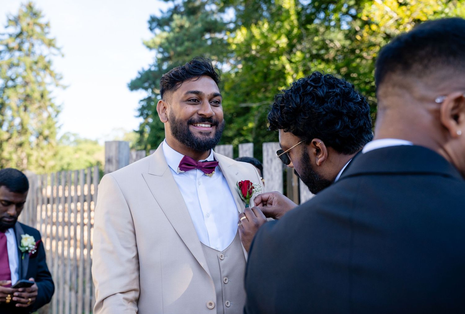 Groom in beige suit with burgundy bow tie smiles as groomsman pins boutonniere at outdoor wedding.