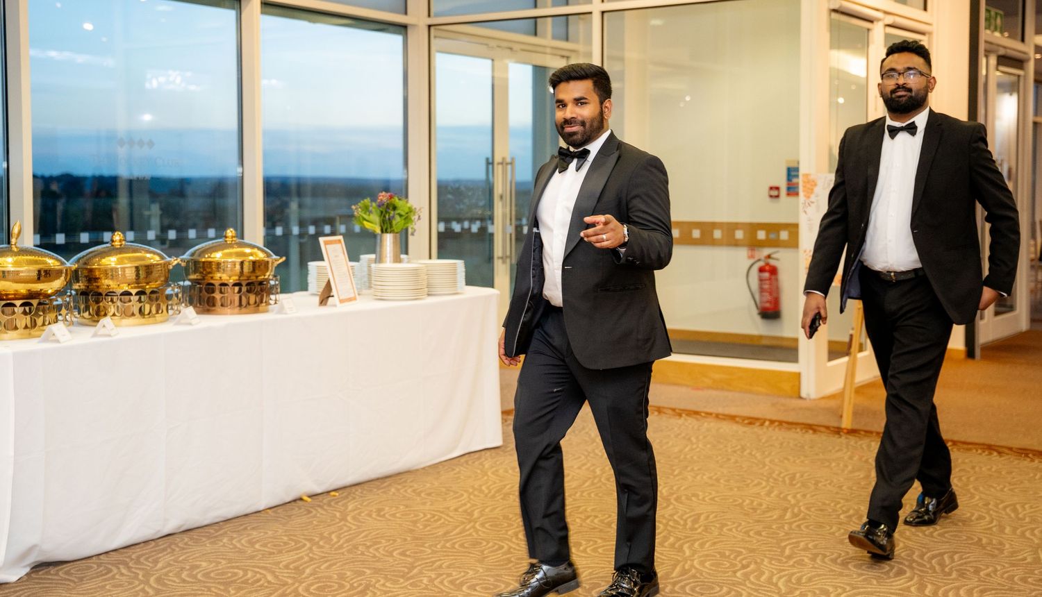 Two men in formal tuxedos walking past a buffet table at a wedding reception.