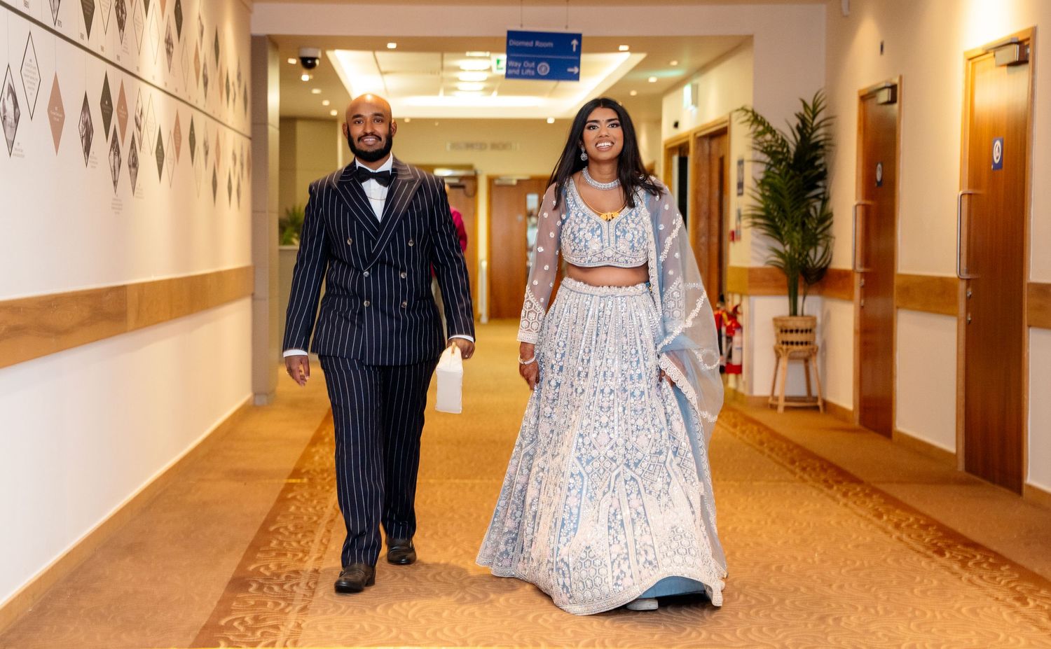Elegant couple walking down a hotel hallway, man in pinstripe suit and woman in silver embroidered lehenga.