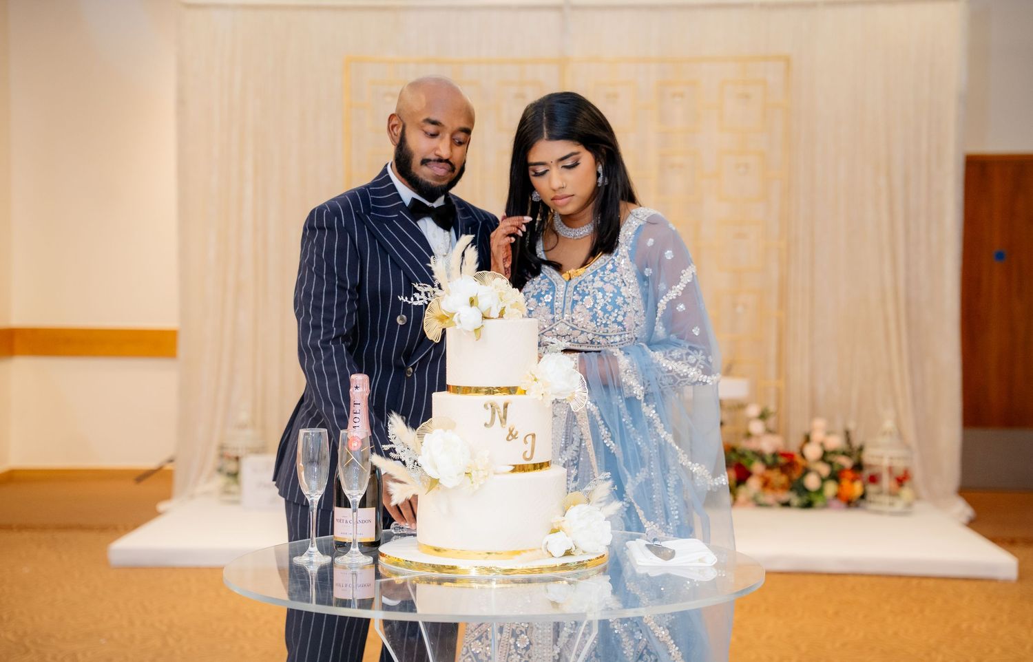 Couple cuts elegant white and gold tiered wedding cake at decorated reception hall.