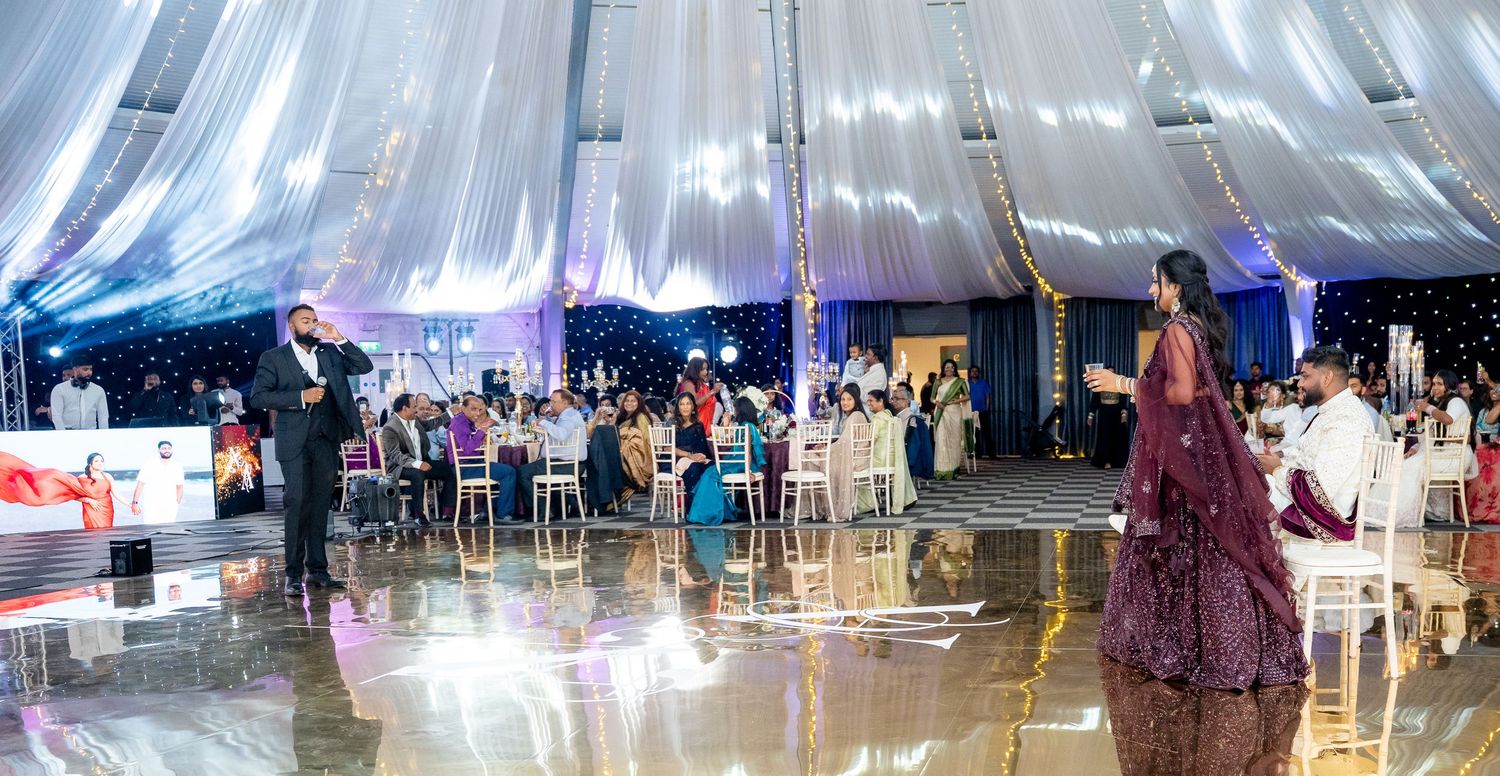 Elegant wedding reception under draped tent with guests seated at tables and dance floor reflecting overhead lighting.