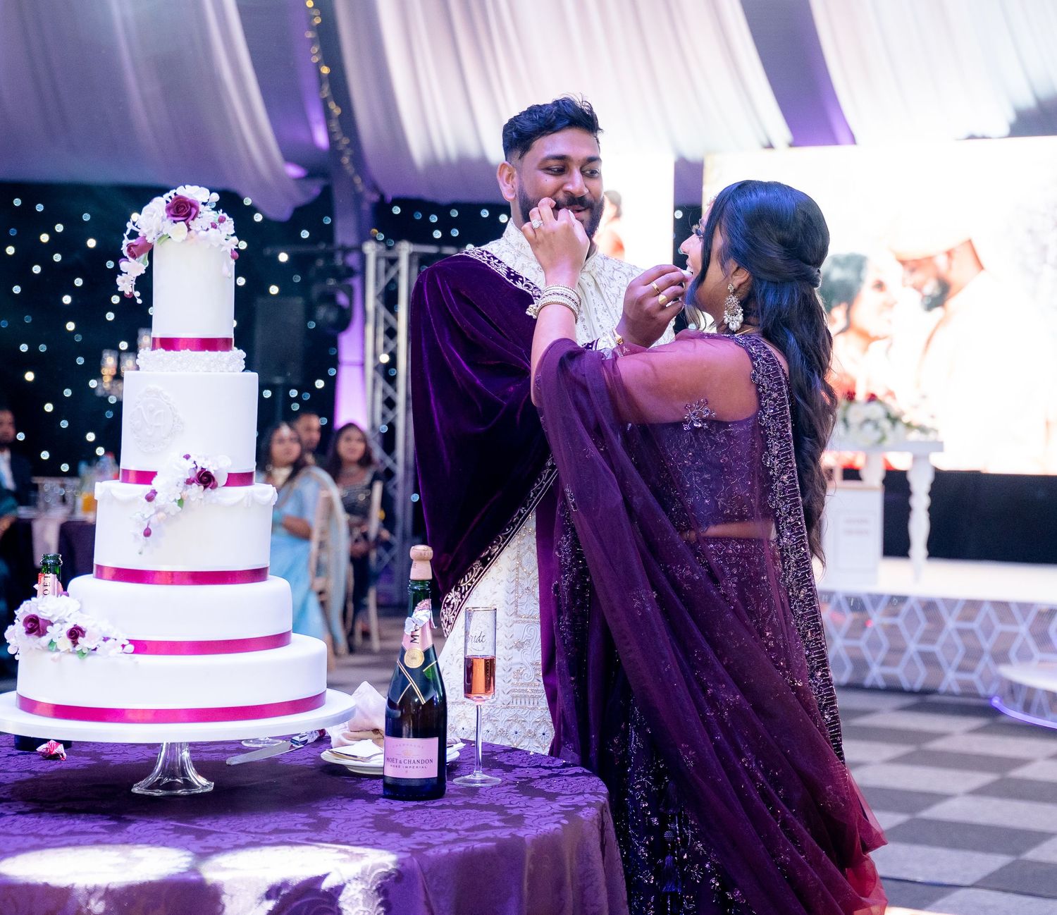 Couple in purple attire cutting a white wedding cake at an elegant reception with champagne and floral decorations.