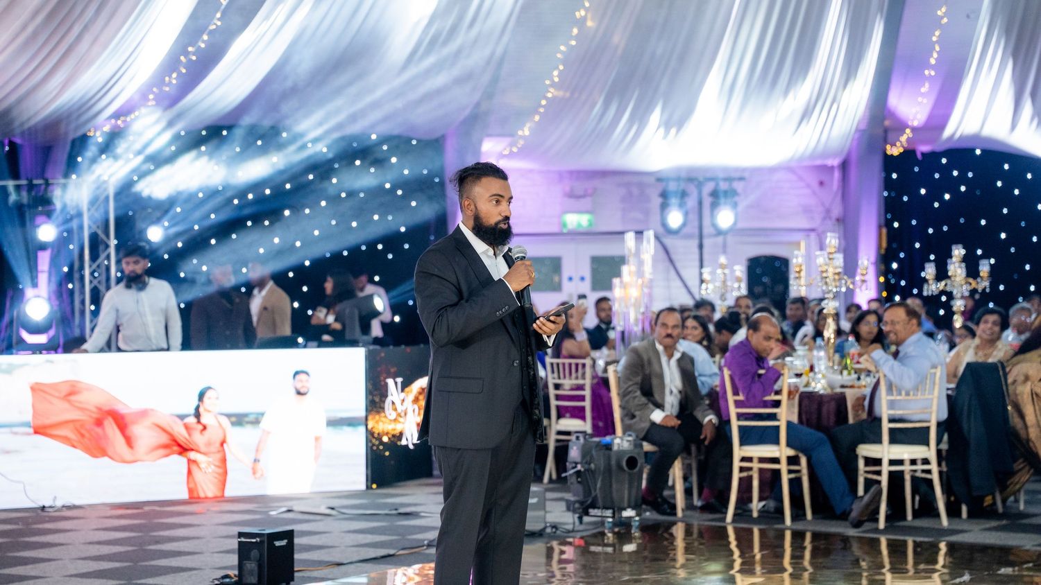 Man in black suit speaks into microphone while hosting an Indian wedding reception in a grand tent.