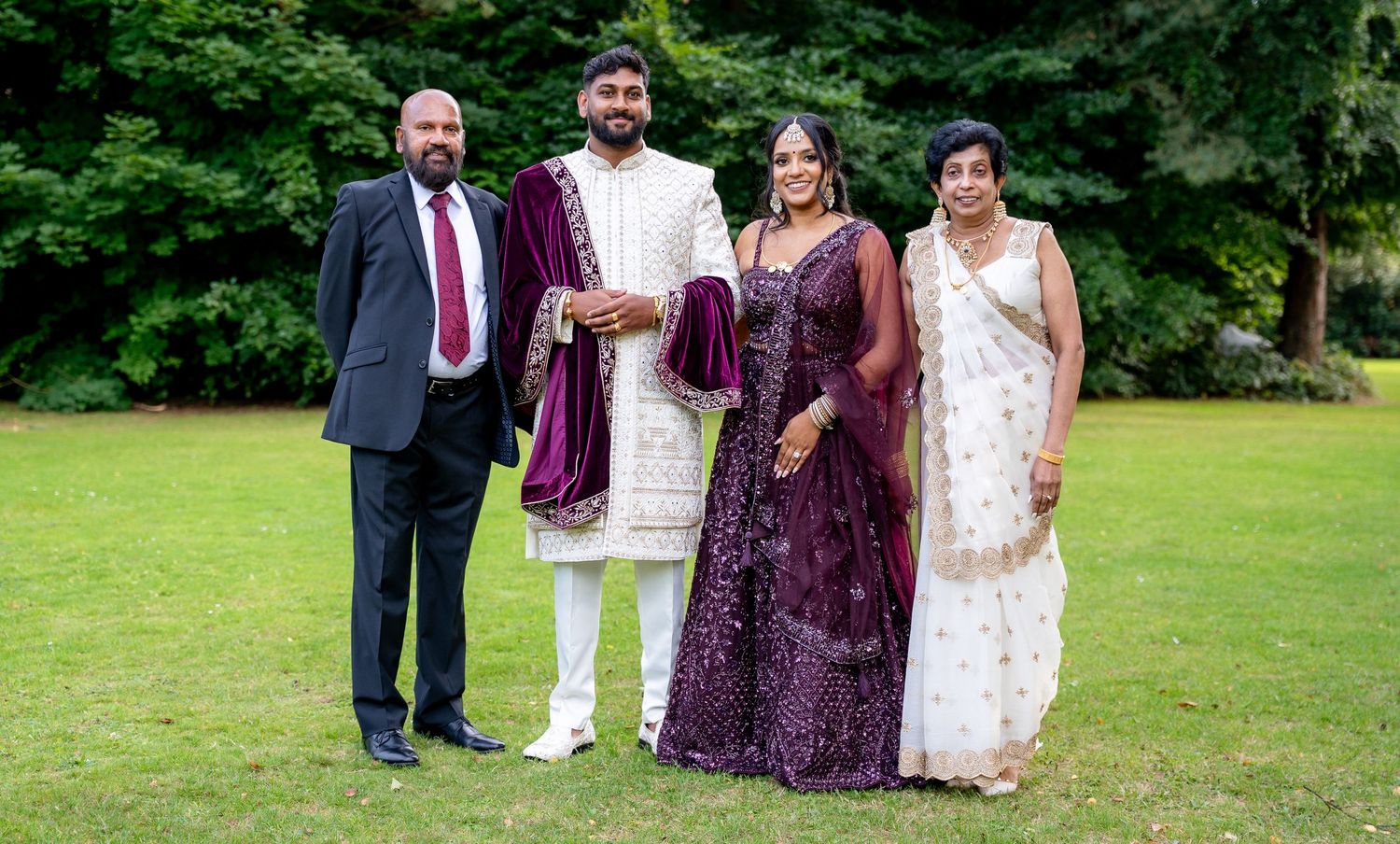 Indian wedding family portrait with four members in traditional attire, purple lehenga and white saree, on green lawn.