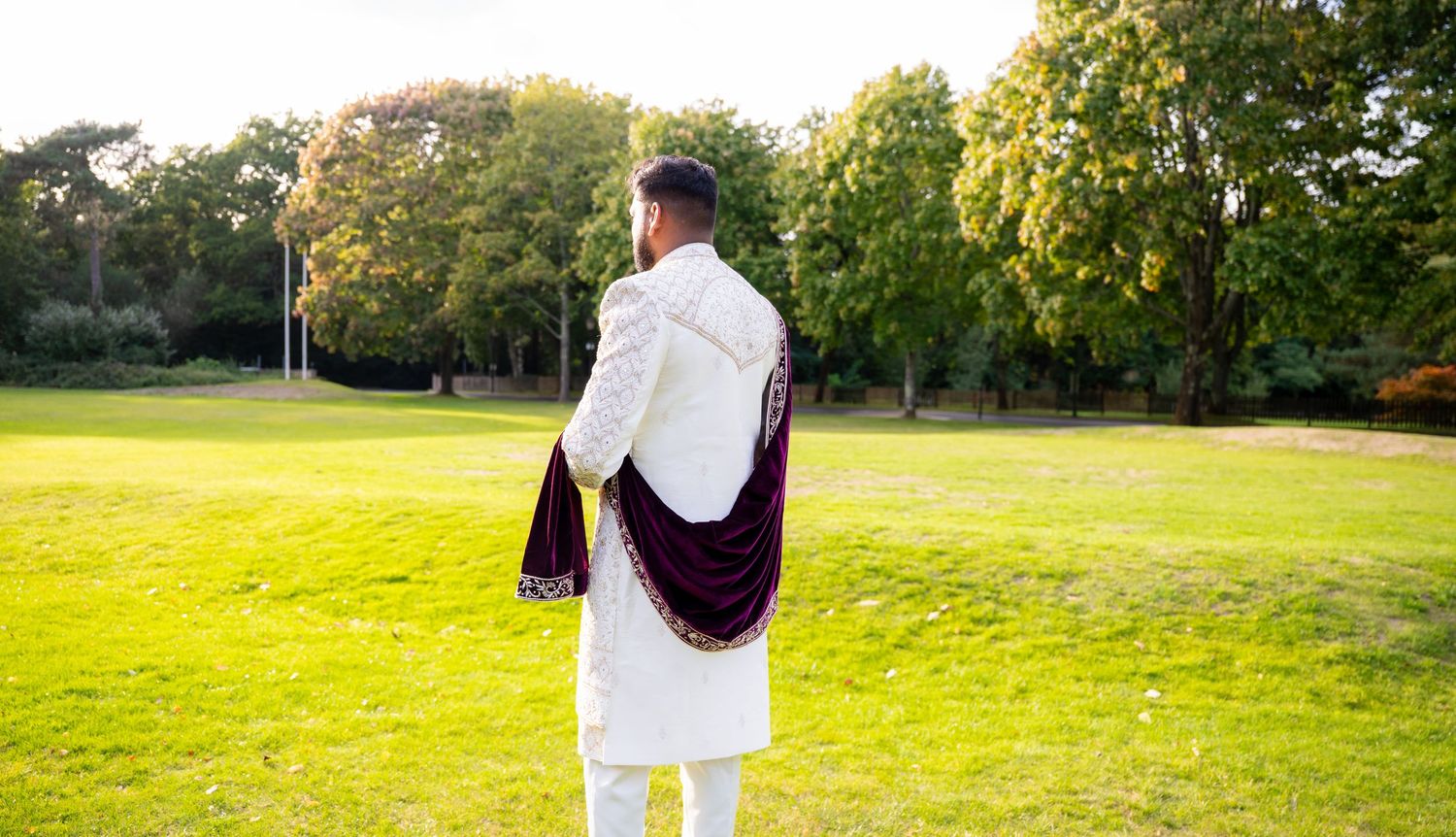 Groom in white sherwani with burgundy velvet dupatta stands in green park, viewed from behind.
