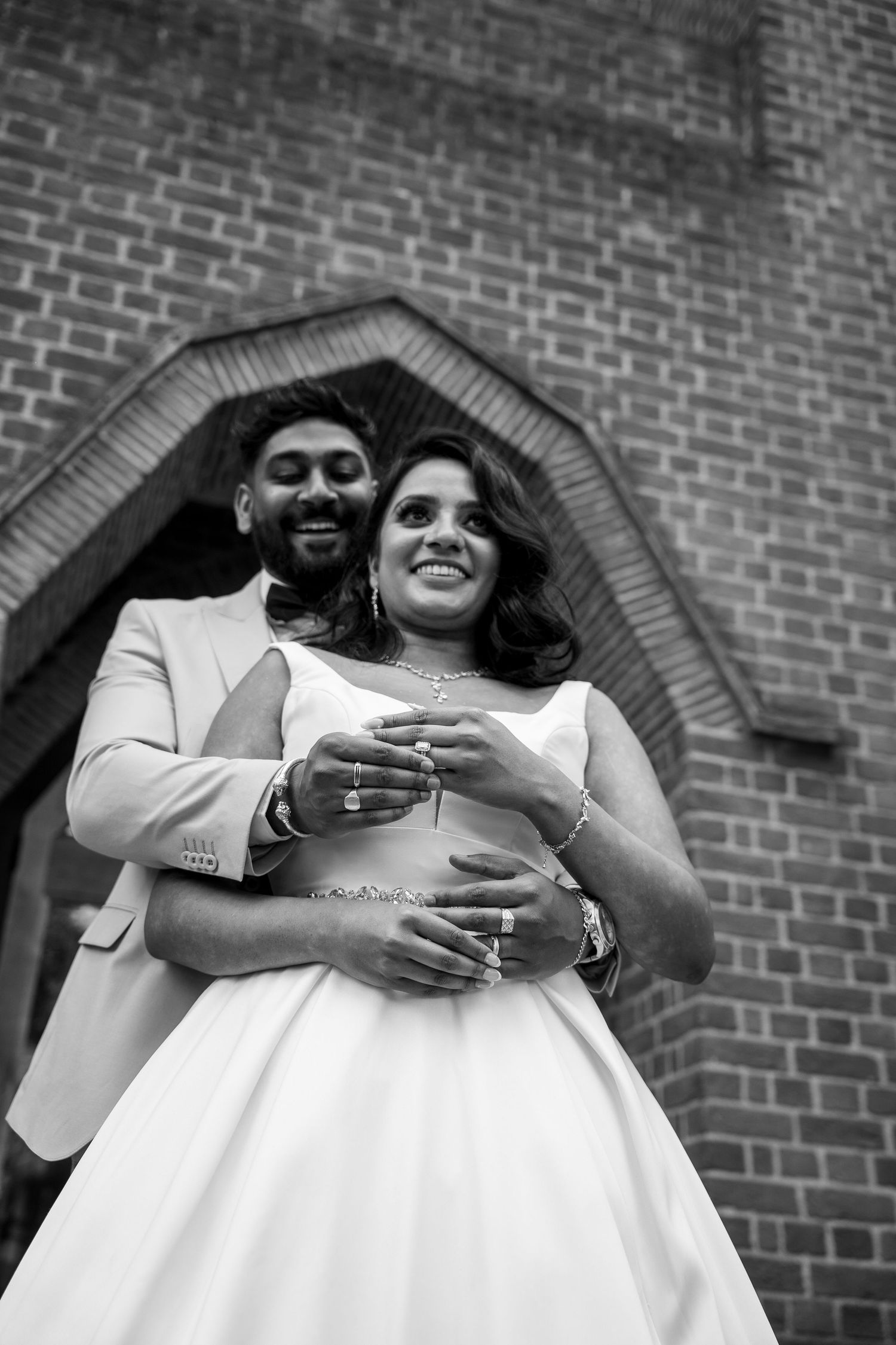 Black and white photo of groom embracing bride in front of brick church archway.