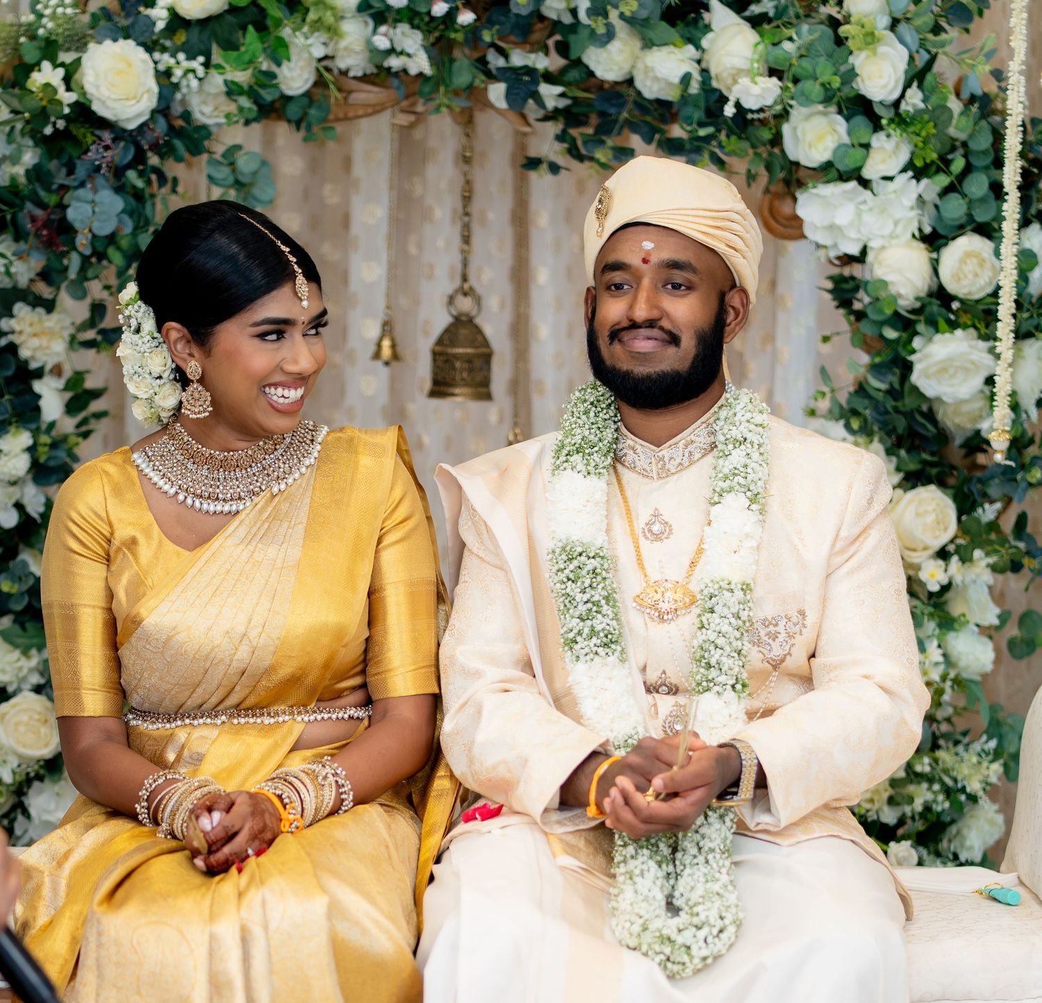 Smiling South Indian bride in gold saree and groom in cream sherwani pose together at floral wedding mandap.
