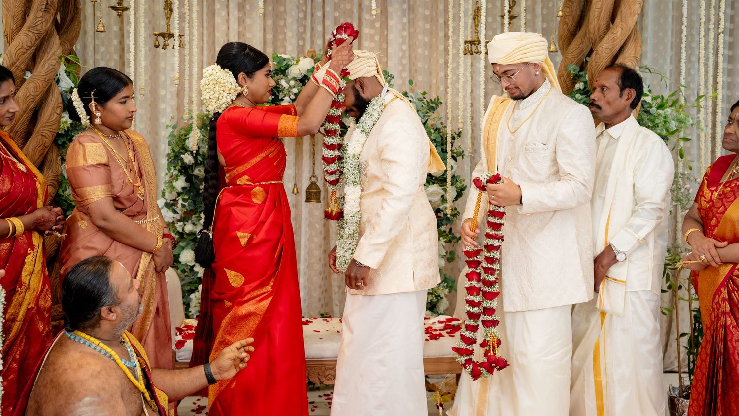 South Indian wedding varmala ceremony with bride in red saree garlanding groom in white sherwani at decorated mandap.