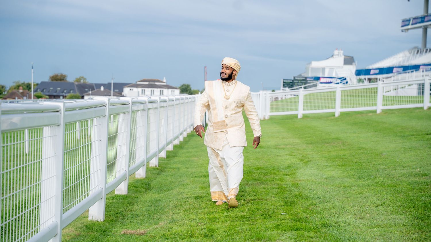 Man in cream suit walking on green grass at a racetrack with white fences and grandstands in background.