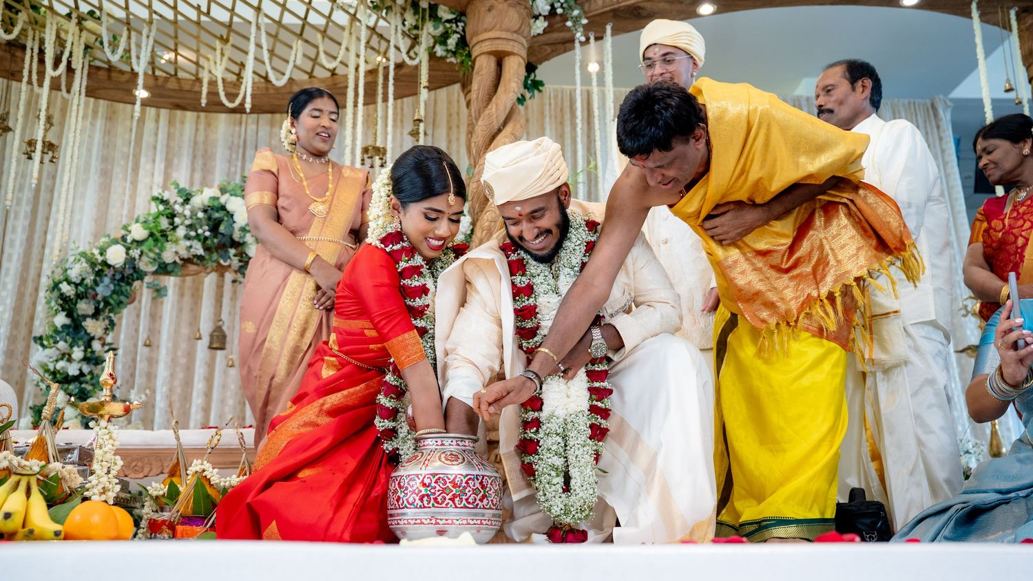 Bride in red saree and groom in white perform a traditional pot ritual during a South Indian wedding.