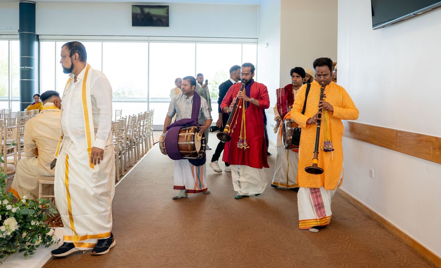 Traditional Indian musicians in yellow and white attire play instruments during a ceremonial procession indoors.