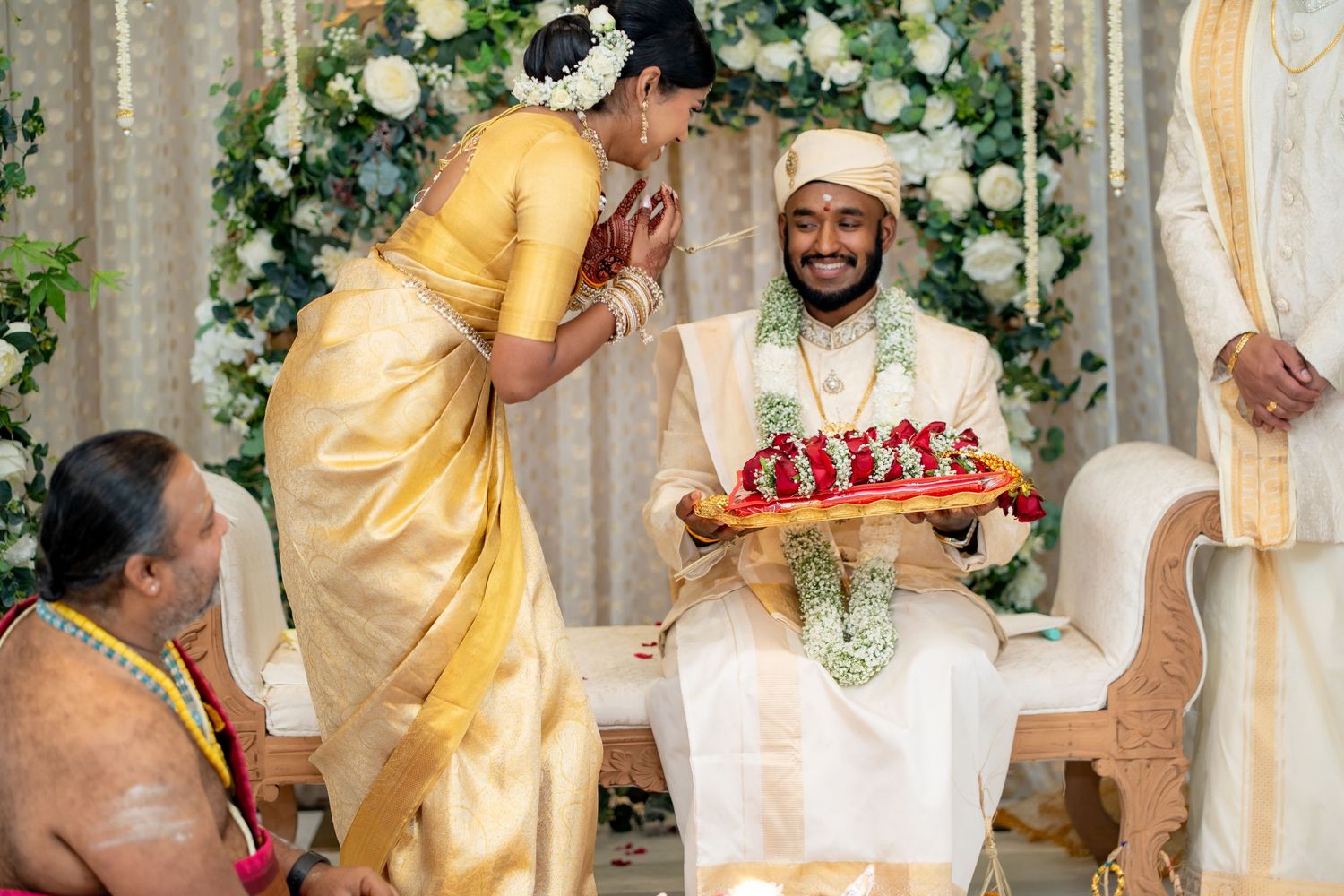 South Indian wedding ceremony with bride in gold saree blessing groom in white sherwani holding floral tray.