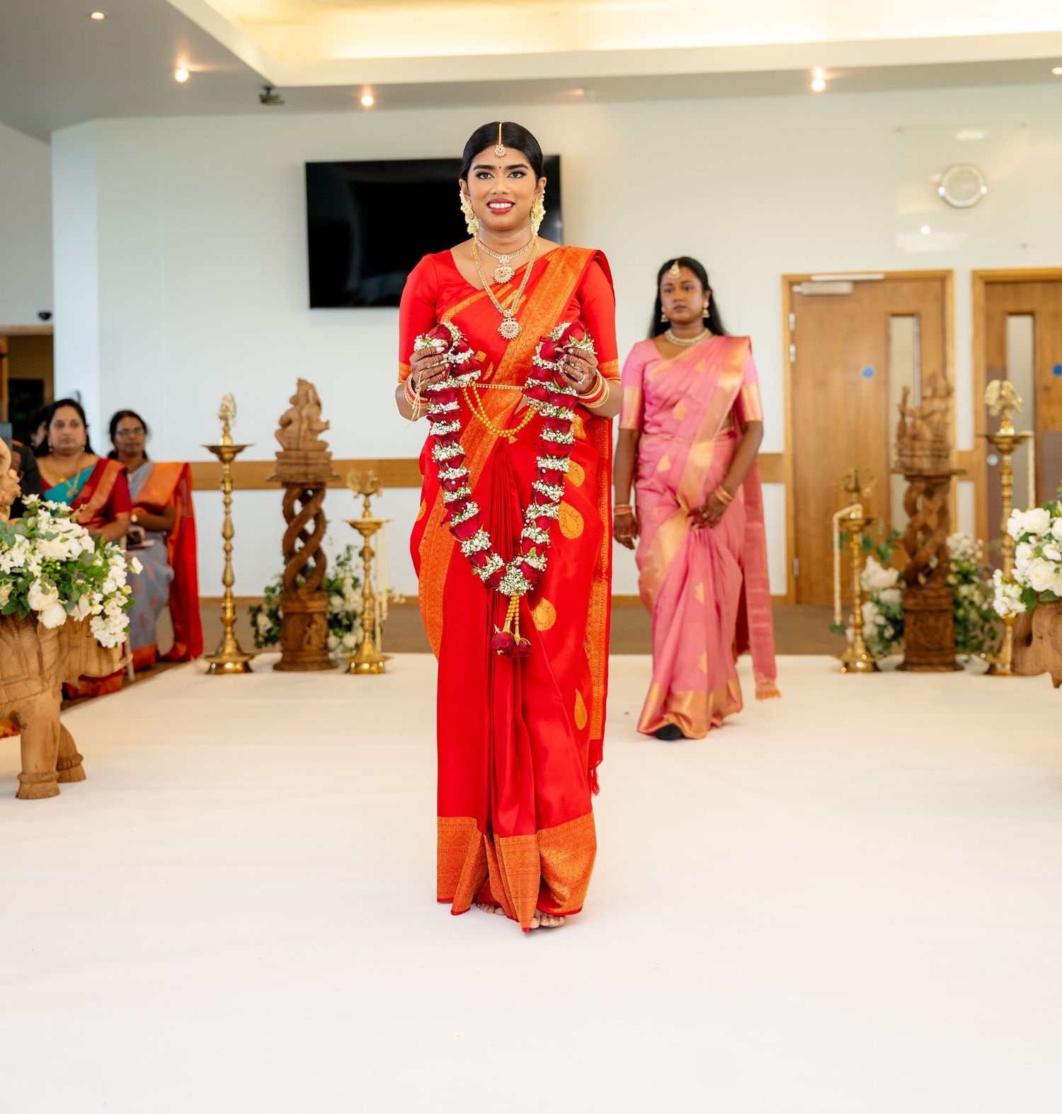 Bride in red saree carrying floral garlands walks down aisle at traditional South Indian wedding ceremony.