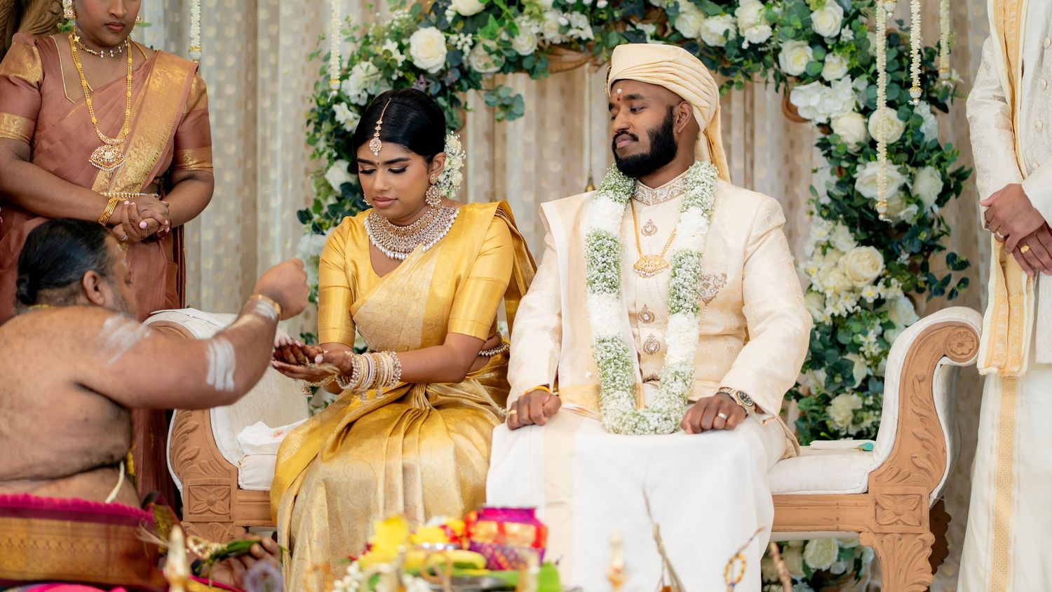 A bride in a golden saree and groom in white sherwani during a traditional South Indian Hindu wedding ceremony.