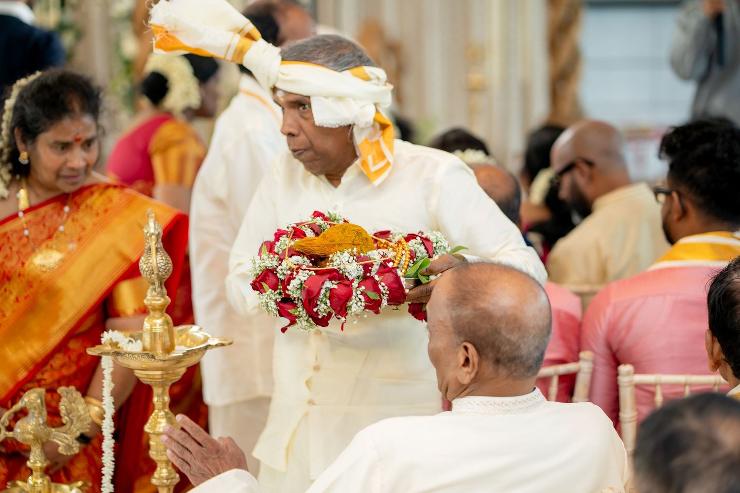 Elder man in white traditional attire presents flower garlands to seated guest at South Indian wedding.