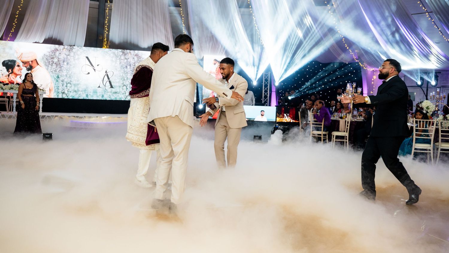 Groomsmen dancing on fog-covered dance floor at elegant wedding reception with purple lighting.