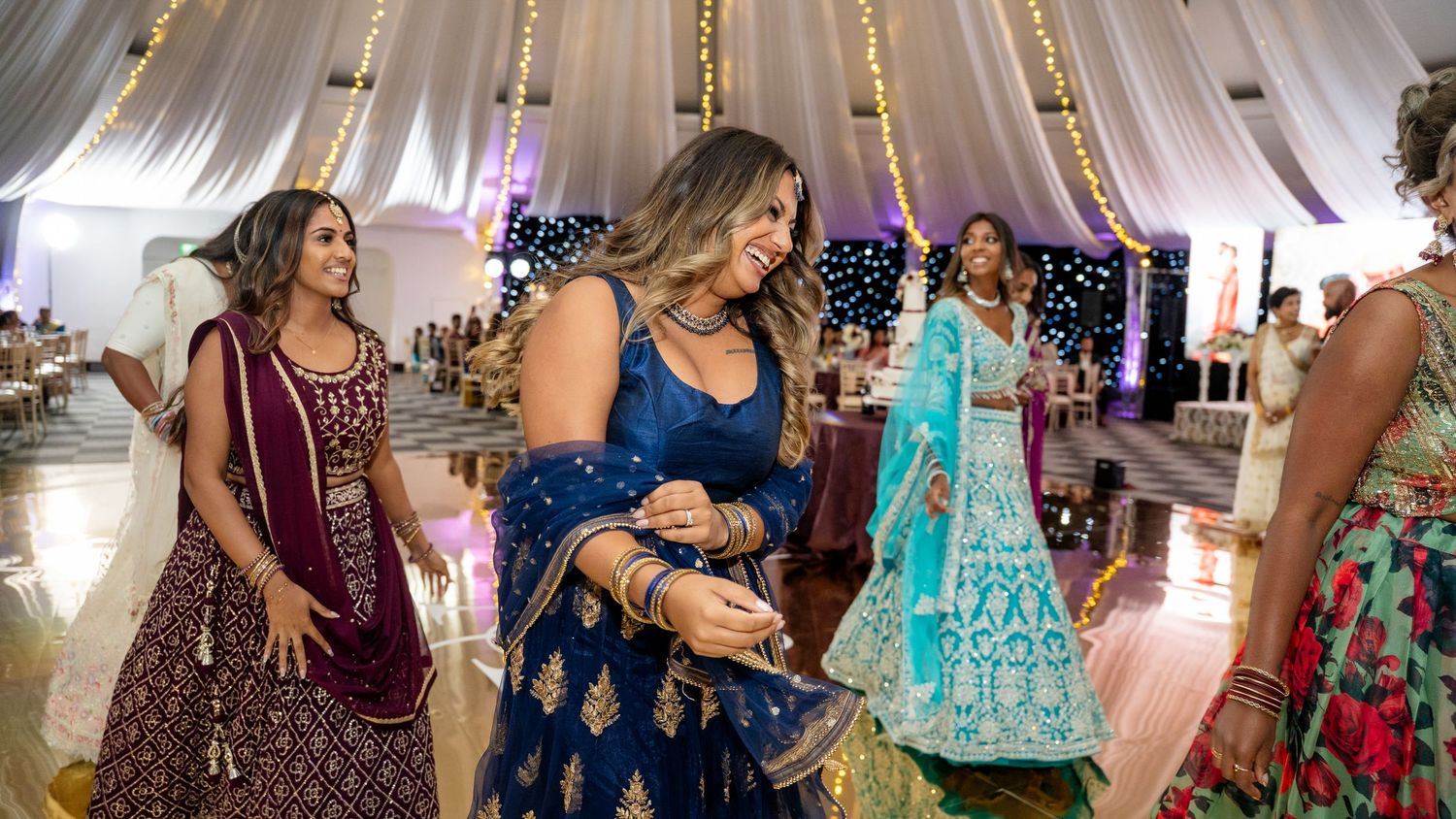 Women in vibrant Indian outfits dance and celebrate together at a warmly lit wedding reception venue.