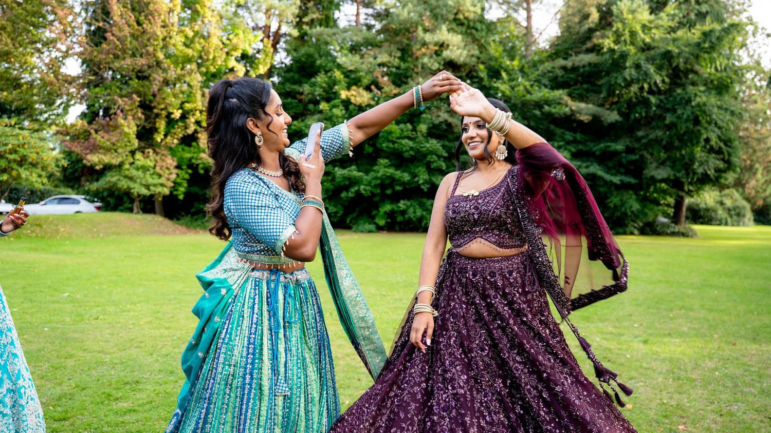 Two women in colorful Indian lehengas spinning and dancing together on a green lawn at a wedding celebration.