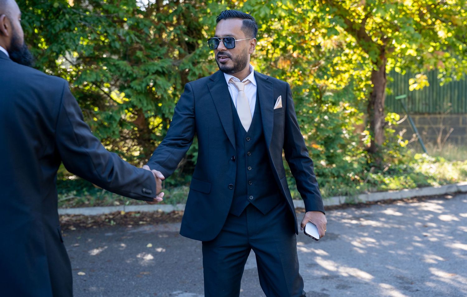 Elegant man in navy three-piece suit and sunglasses shaking hands outdoors.