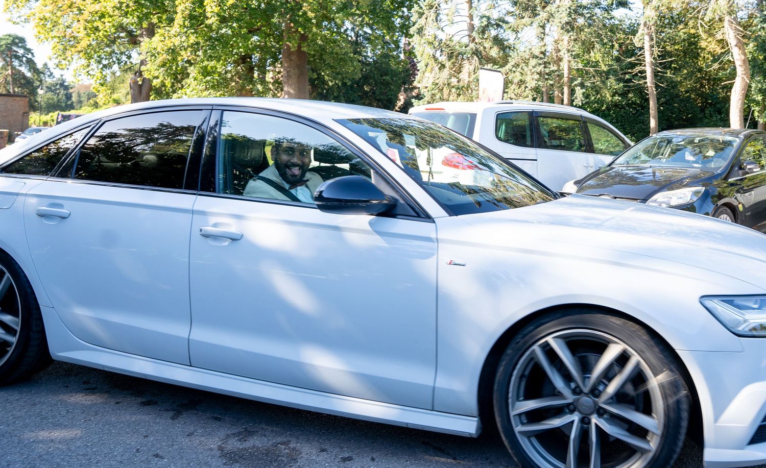 White Audi sedan parked among trees in a sunlit outdoor parking area at a wedding venue.