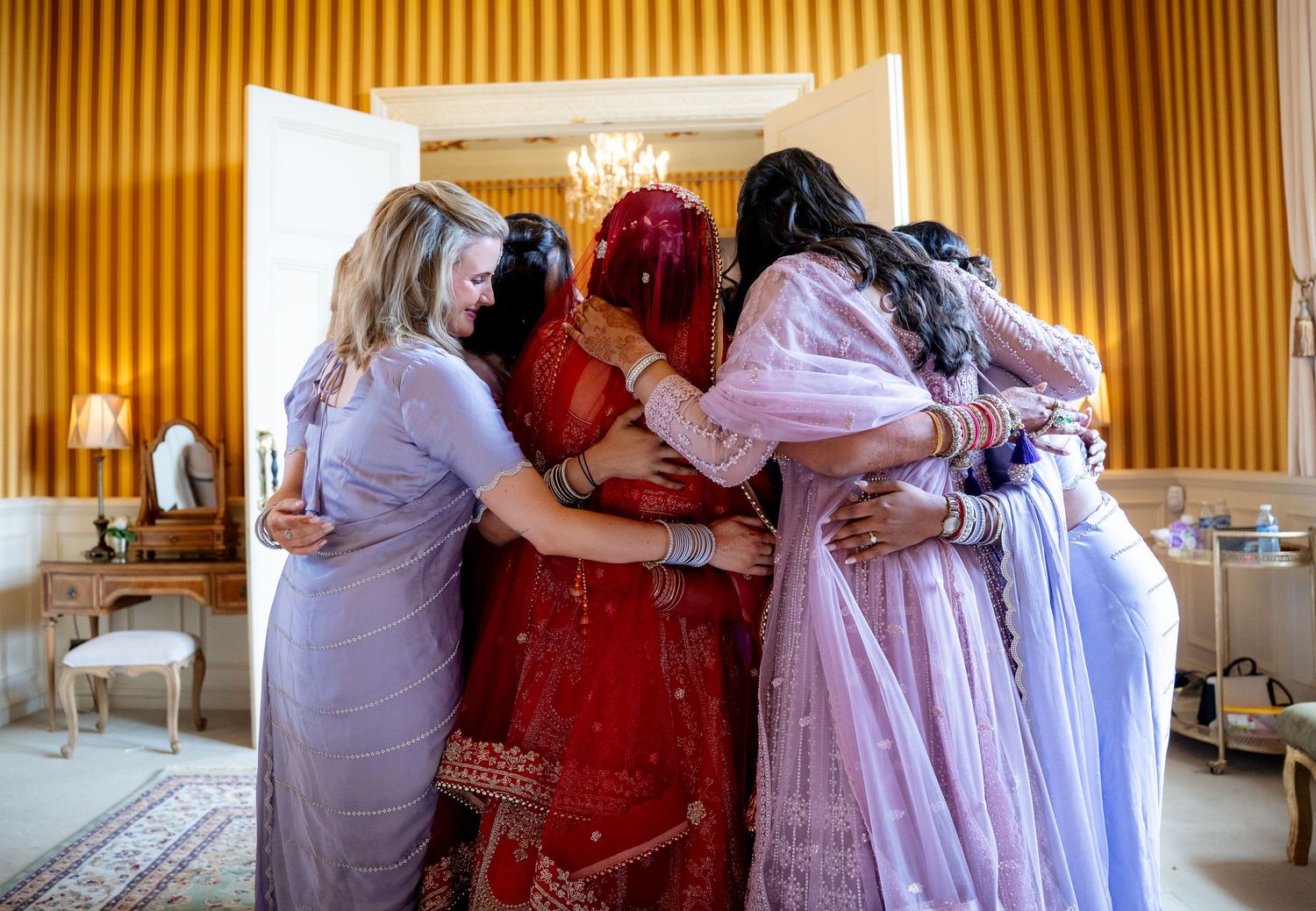 Indian bride in red lehenga shares emotional group hug with bridesmaids in lavender dresses.