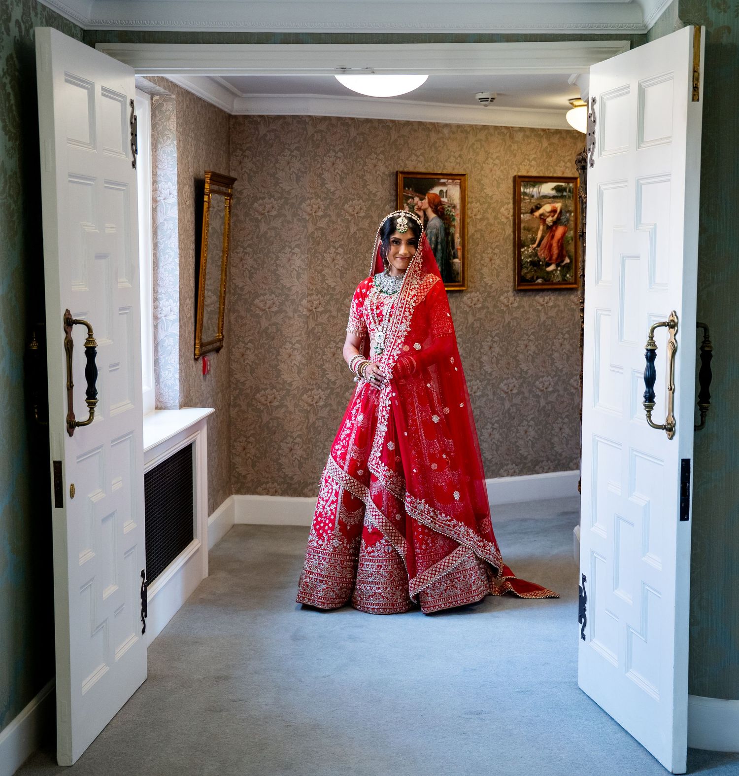 Indian bride in red bridal lehenga standing elegantly between open white doors in ornate room.