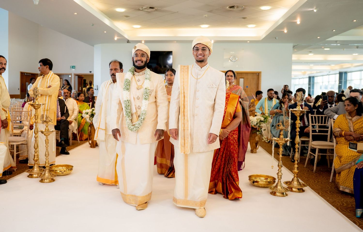 Two grooms in white traditional attire walking down the aisle at a South Indian wedding.