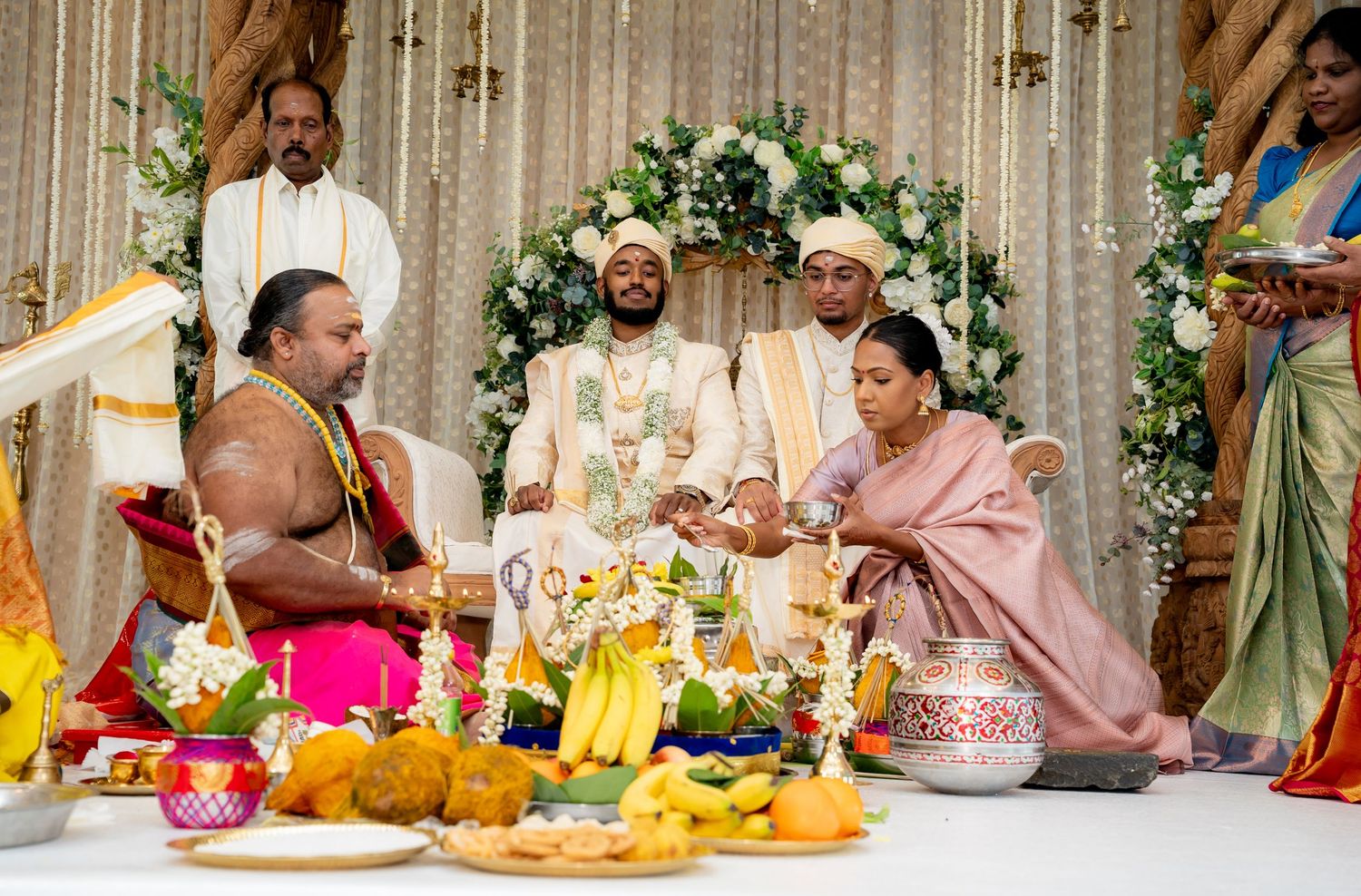 Traditional Hindu wedding ceremony with priest, couple, and family gathered around ritual offerings and decorations.