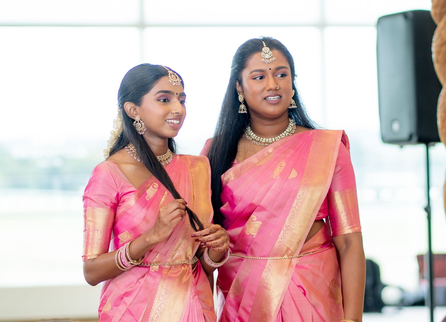 Two young women in matching pink silk sarees with gold jewelry at a South Indian wedding celebration.