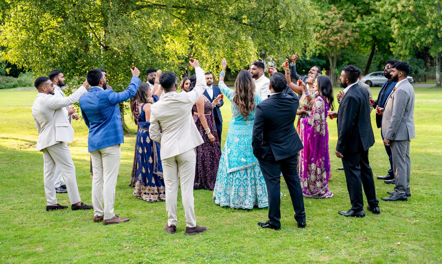 Large group of wedding guests in traditional Indian attire celebrating outdoors with raised hands on a green lawn.