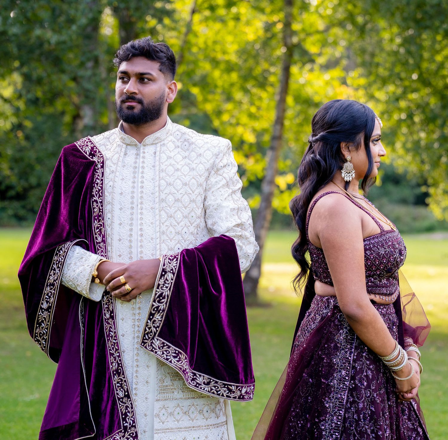 Indian couple in coordinating purple and white traditional wedding outfits posing outdoors in garden.