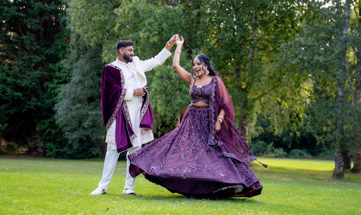 Couple in traditional Indian wedding attire dance joyfully outdoors with lush green trees in the background.