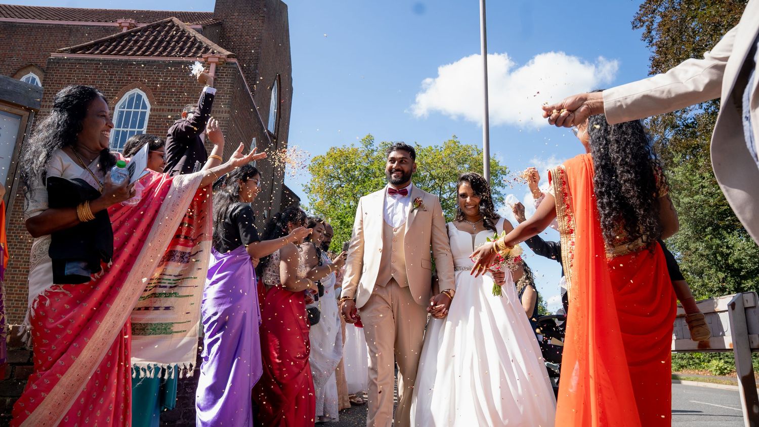 Joyful couple walks through celebratory send-off with guests in colorful saris waving sparklers outside a church.