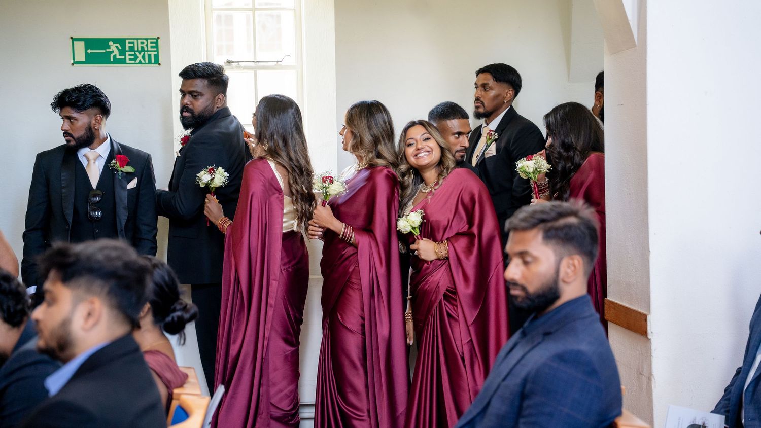 Bridesmaids in burgundy dresses and groomsmen in black suits gather in church foyer before ceremony.