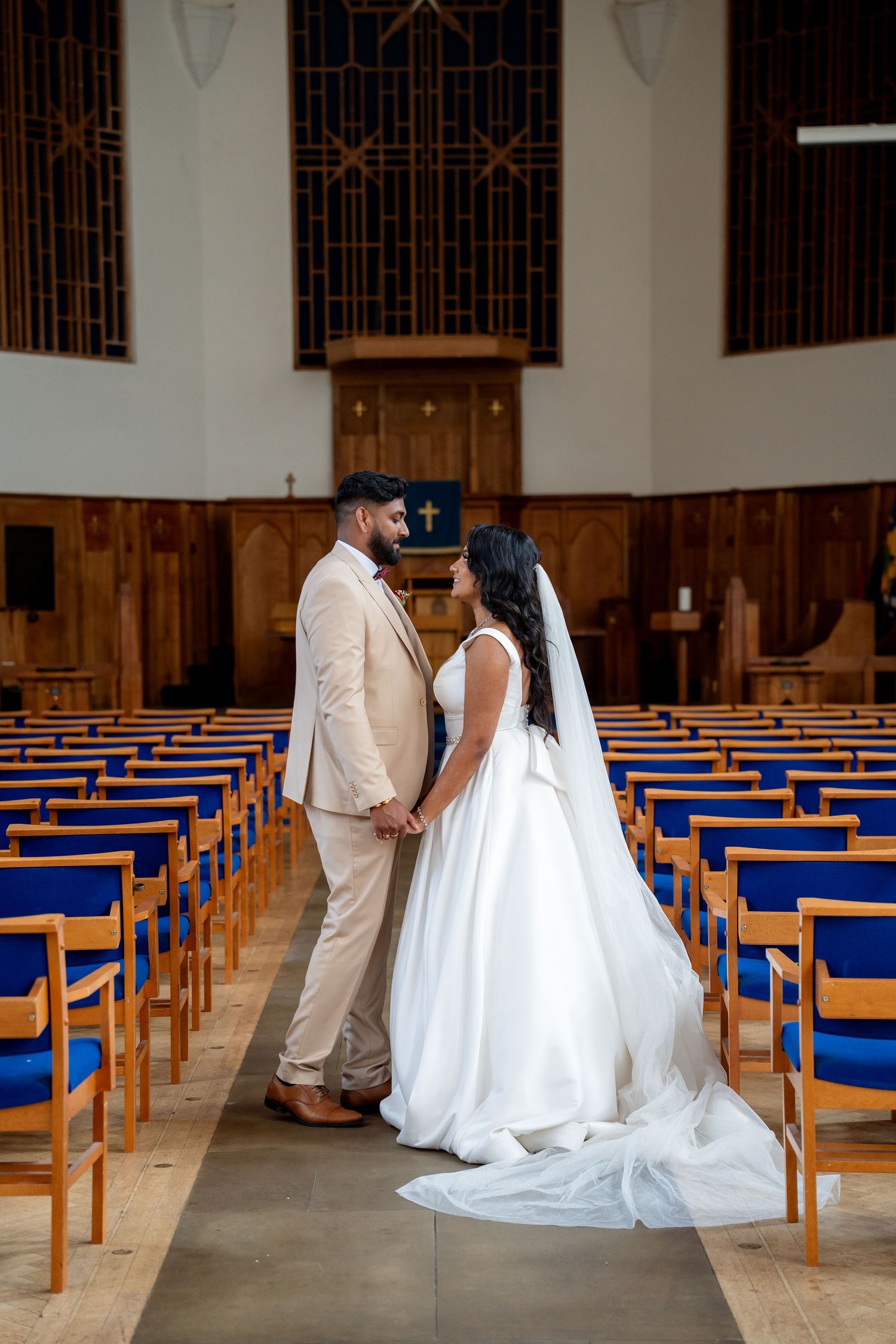 Bride and groom hold hands in a church aisle lined with blue pews, bride wearing a long veil and white gown.