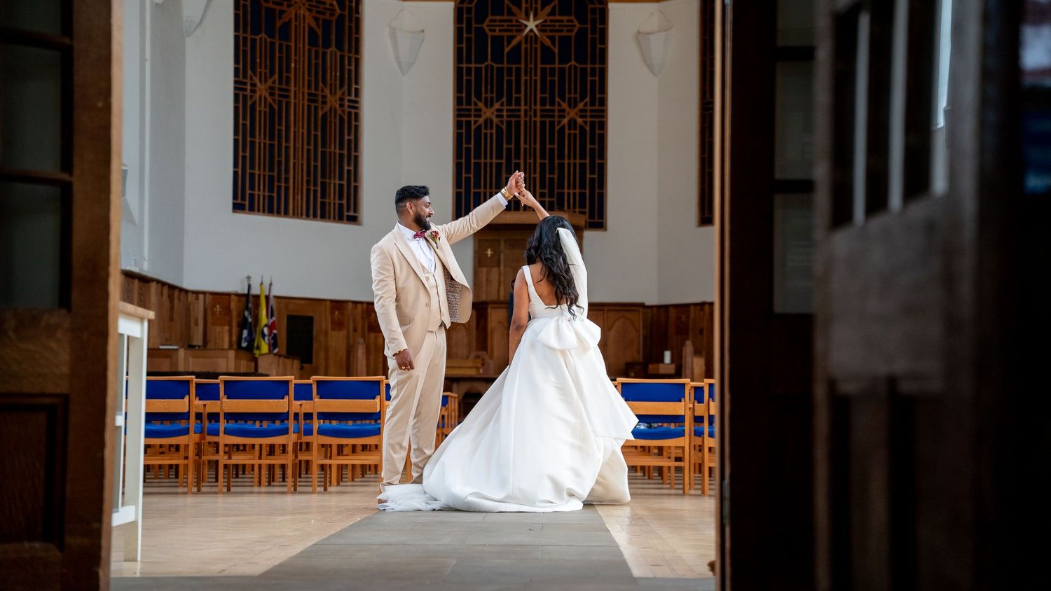 Newlyweds share a tender dance moment in an elegant church, groom in tan suit spinning bride in flowing white dress.