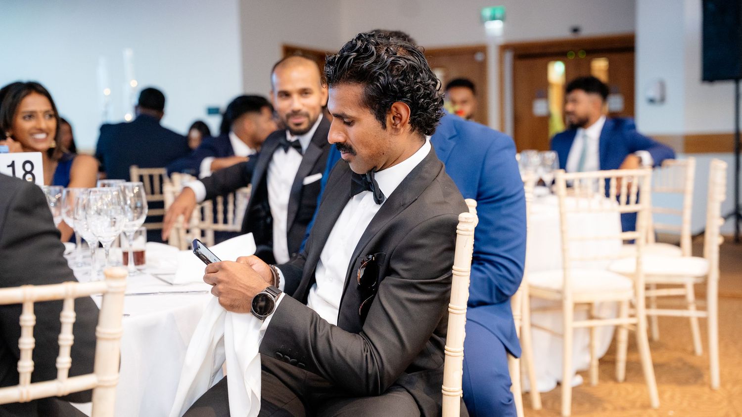 Man in tuxedo checking phone at a formal wedding reception dinner table.