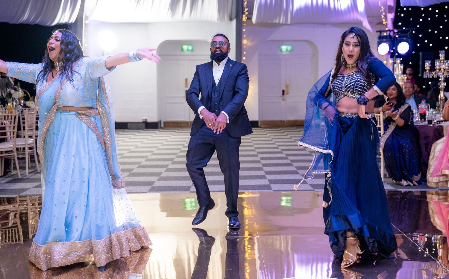 Man in navy suit dancing between two women in blue Indian wedding attire at reception.