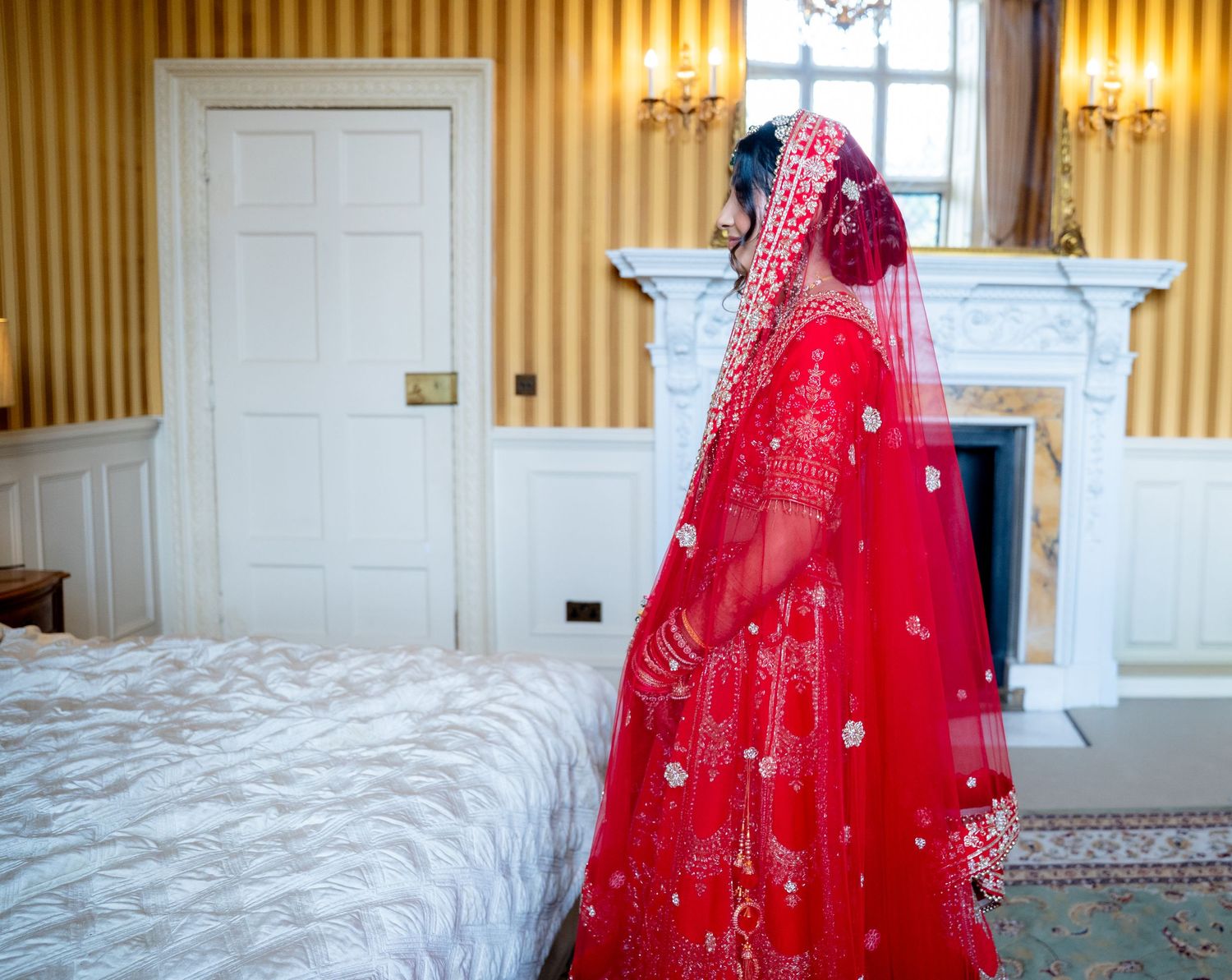 Indian bride in red lehenga with embroidered dupatta stands by fireplace in elegant room.