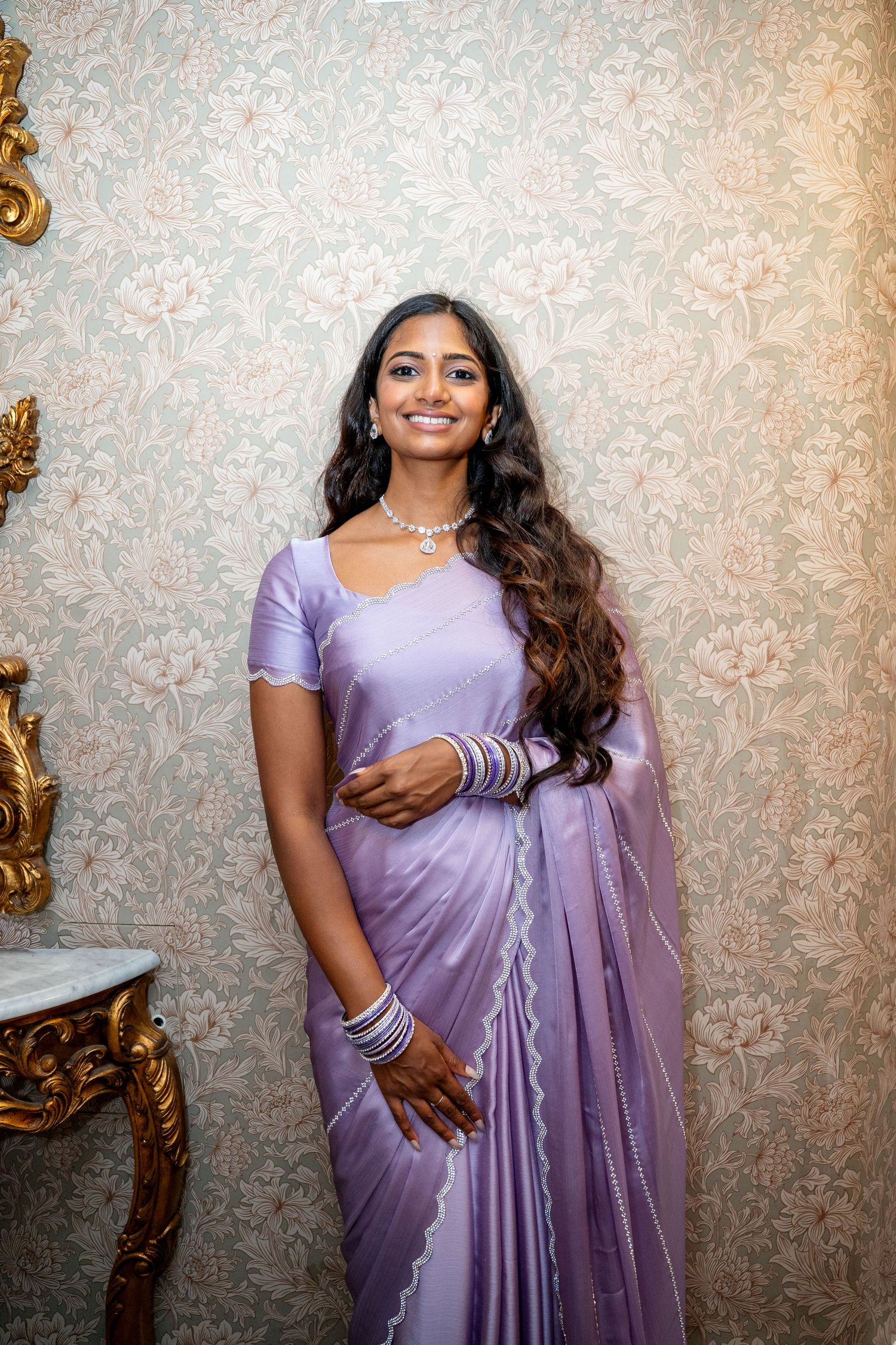 Young woman in lavender silk saree with silver bangles and necklace against floral wallpaper.
