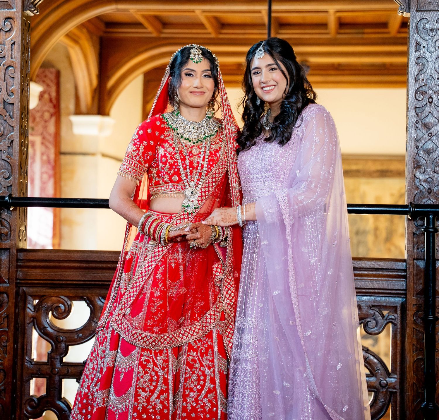 Two women in traditional Indian attire, one in red bridal lehenga and one in lavender salwar, posing together.