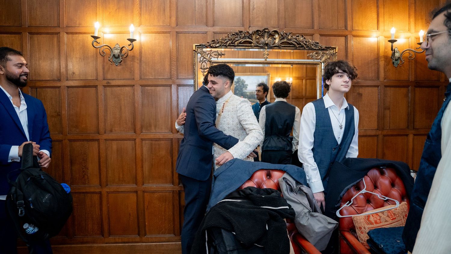 Men in formal attire gather in a wood-paneled room, socializing near a mirror and a stroller at an elegant event.