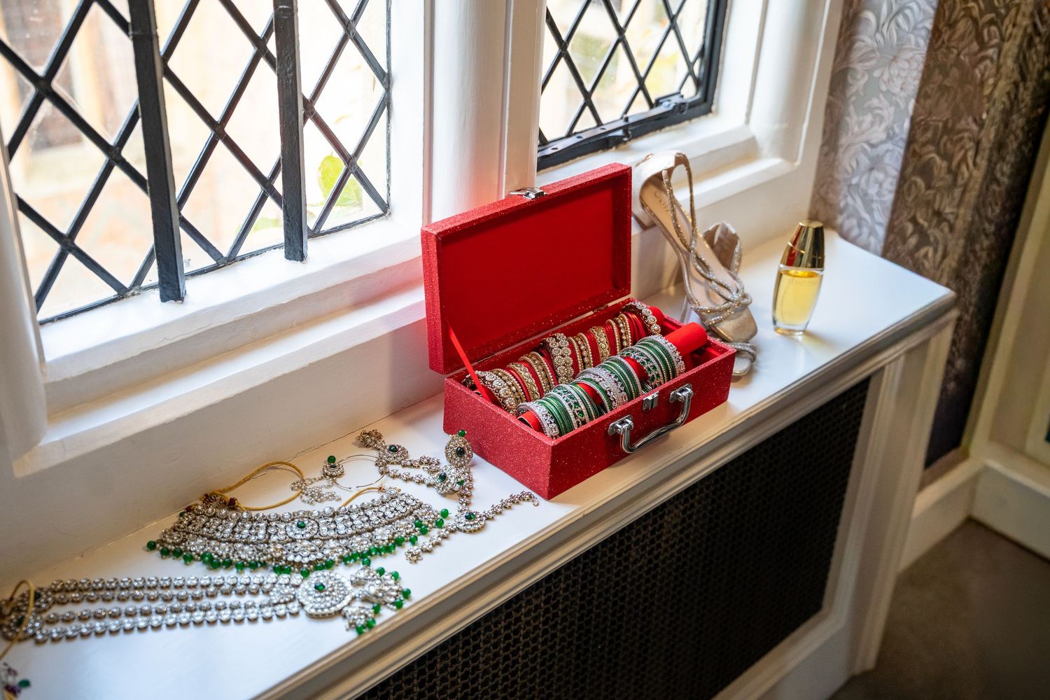 Red jewelry box with bangles and bridal accessories displayed on windowsill with leaded glass windows.