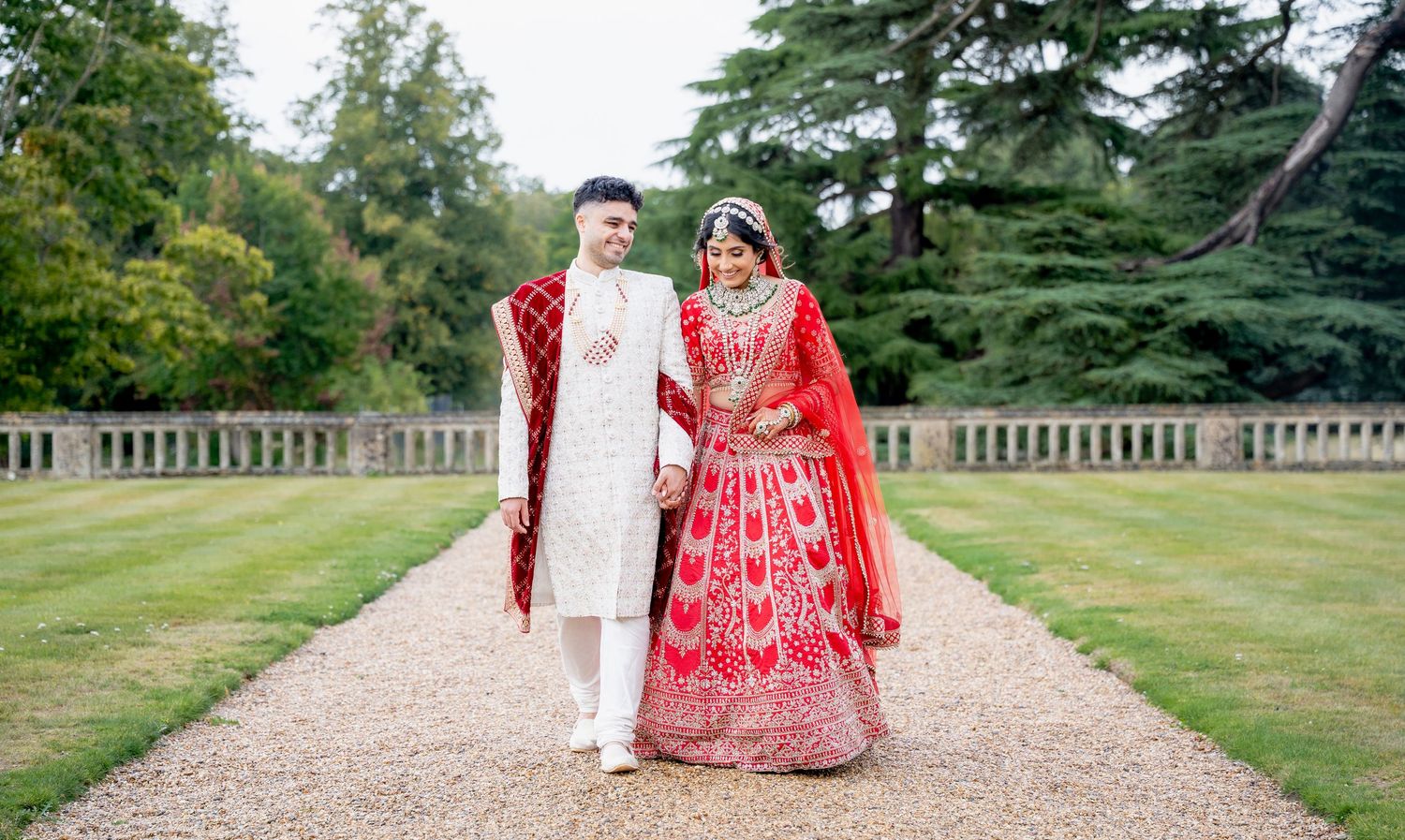 Indian bride and groom in vibrant red and white wedding attire walk hand in hand along a garden path.